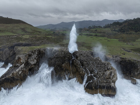 Aerial view of the Bufones de Pria where seawater shoots upwards with great force against a backdrop of green hills, Llanes, Asturias, Spain.