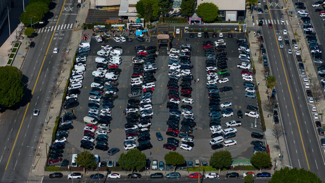 Aerial view of a busy parking lot filled with cars, nestled between wide streets and tall buildings, casting stark shadows under the sun, Los Angeles, California, United States.