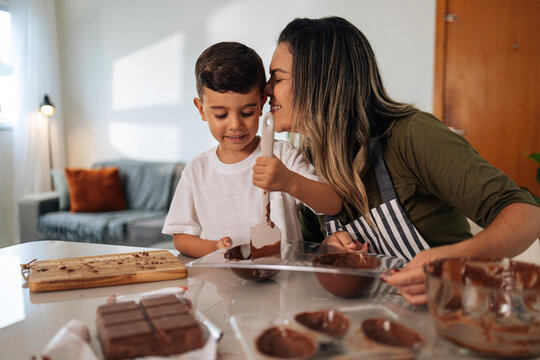 Brazilian mother helping child fill molds with chocolate to make Easter eggs and bunny candies, showing handmade dessert tradition and family celebration indoors.