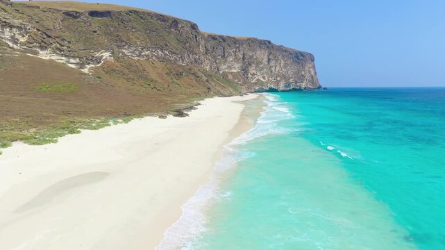 Aerial view of a white sand beach and turquoise ocean water along steep rocky cliffs at Socotra Island, Yemen.