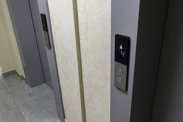 Two modern elevator doors with digital panels showing floor number «4» and up arrow. Light beige walls, grey tiled floor, sleek metallic design, two call buttons per panel. © evgeniykleymenov
