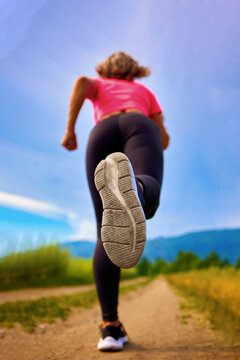 An athletic woman is sprinting along a field road, framed by a breathtaking mountain backdrop and a clear blue sky. The scene captures the essence of determination and the beauty of nature.