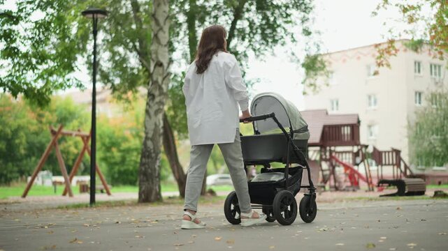 Mother walking stroller toward playground, leafy trees and residential buildings in background, soft daylight, attentive pace, protective posture, emphasis on future and bonding, tranquil urban