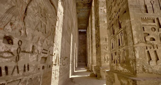 Medinet Habu temple colonnade with hieroglyphic inscriptions and relief carvings in Luxor Egypt, massive sandstone columns with papyrus capitals under dramatic blue sky.
