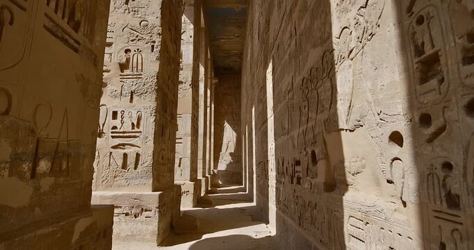 Medinet Habu temple colonnade with hieroglyphic inscriptions and relief carvings in Luxor Egypt, massive sandstone columns with papyrus capitals under dramatic blue sky.
