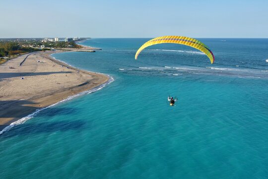Aerial view of a paraglider soaring above the turquoise water near a sandy beach and buildings along the coast, Jupiter, Florida, United States.