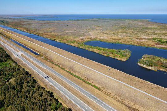 Aerial view of a highway and a canal dissecting the landscape, bordered by deep green trees and expansive wetlands near Lake Okeechobee, Lake Harbor, Florida, United States.