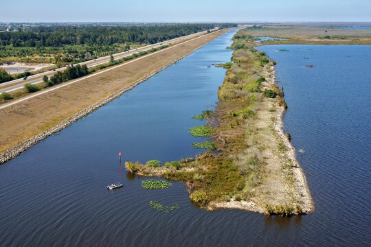 Aerial view of a tranquil canal reflecting the clear sky, bordered by a levee and lush vegetation, contrasting with the deep blue waters of Lake Okeechobee, Lake Harbor, Florida, United States.