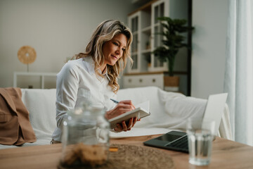 Woman working remotely from home office, writing notes in notebook while using laptop on sofa
