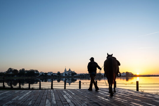 Cowboy Standing With Mustang Beside Lake At Sunrise