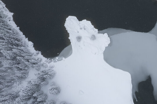 Aerial view of a snow-laden forest kissing the icy embrace of Lac de Taney, a stark contrast of textures under a winter sky, Vouvry, Valais, Switzerland.