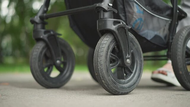 Family strolling outdoors with kids in tow. Parent pushing pram along path with leaves and pavement. Caregiver briskly pushes stroller over pavement surrounded by fallen leaves and nature