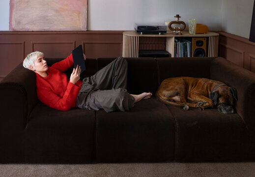 Person In Red Sweater Reading Tablet On Couch While Dog Sleeps Beside
