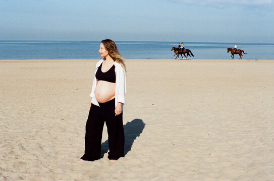 Pregnant woman standing on sandy beach with horses in background 