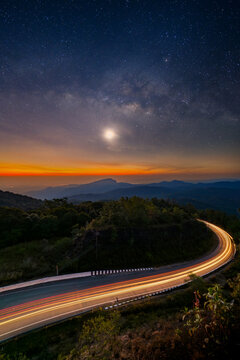 Long exposure of car light trails on a winding road at Doi Inthanon, Thailand. Stunning night landscape with the Milky Way galaxy, moon, and orange twilight glow over mountain ranges.
