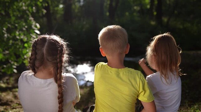 children sit on the banks of the river. happy family childhood dream concept lifestyle. a group of little children sit together by the river and talk. kids spend time outdoors in nature in the forest
