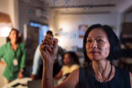 Woman writes math equations on glass, colleagues in background.