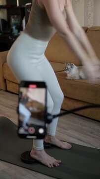 Home fitness coach filming squat exercise on smartphone in living room. Athletic woman performing workout on mat, producing training tutorial, emphasizing technique, leg power and buttock muscles