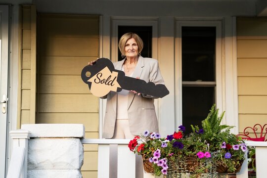 Senior realtor holding sold key sign on house porch