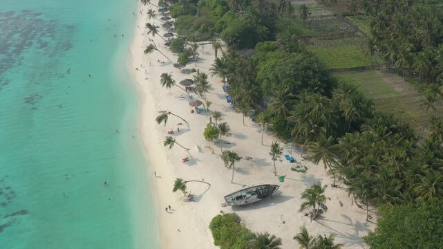 Aerial view of a white sand beach on Thoddoo island with a wrecked wooden boat, lush green palm trees, tourists relaxing, and clear turquoise ocean water.