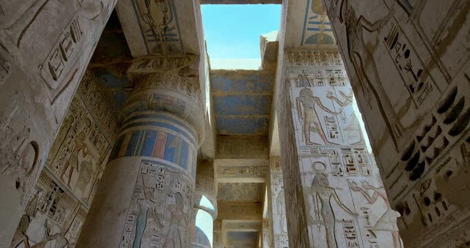 Medinet Habu temple colonnade with hieroglyphic inscriptions and relief carvings in Luxor Egypt, massive sandstone columns with papyrus capitals under dramatic blue sky. Mortuary Temple of Ramesses II