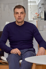 Man sitting in armchair at home, looking surprised or concerned, indoor lifestyle portrait with neutral background and natural light