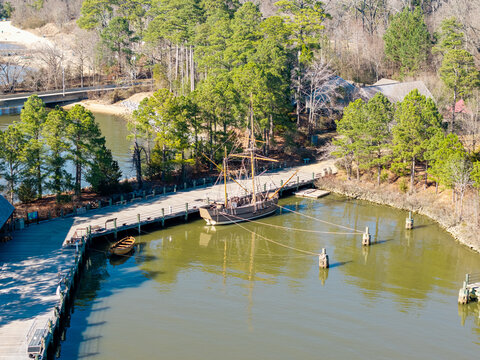Colonial-era merchant ship replica, the Godspeed, docked at the wooden pier of the Jamestown Settlement living history museum on the James River. Aerial view.
