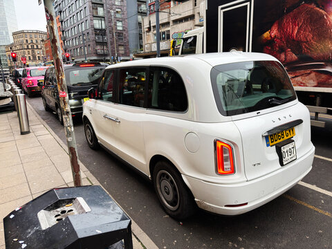 White LEVC TX taxi on a busy Manchester street amid traffic and tall city buildings