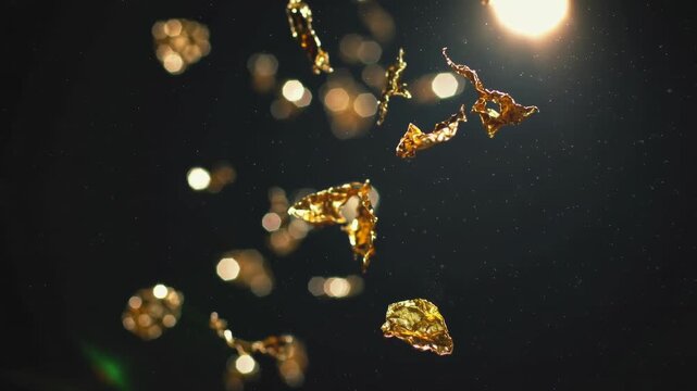 Ultra-realistic luxury abstract video featuring ultra-thin gold leaf fragments floating slowly in the air against a dark background. Warm spotlight illumination creates soft reflections