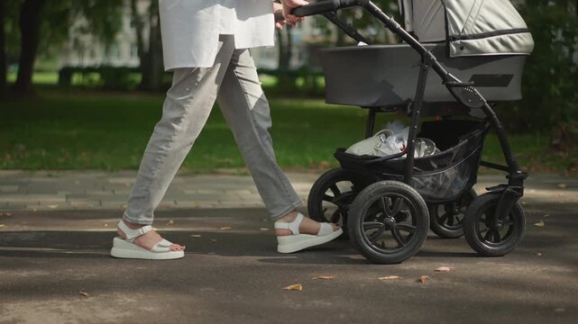 Closeup legs pushing stroller in park young caucasian mother in casual trousers and sneakers bassinet pram under soft morning sunlight lowangle framing calm routine and gentle motion lifestyle decree