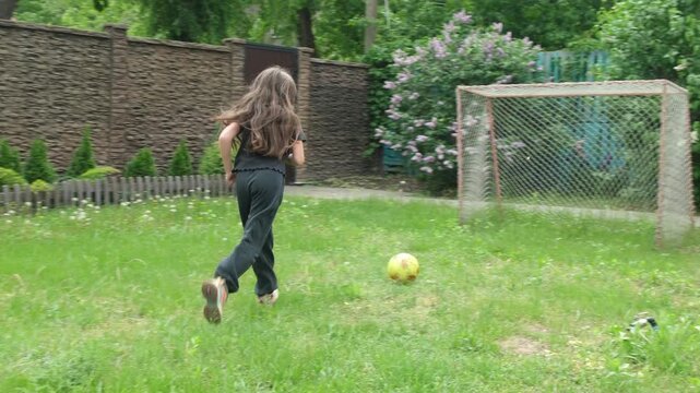 Active little girl with long hair kicking a soccer ball into a goal net on the green grass of a home backyard during a sunny day, showcasing a healthy and fun childhood lifestyle