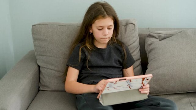 Annoyed little girl sitting on a couch, becoming upset and making a facepalm gesture after losing an online game on her tablet, showing disappointment and negative emotions