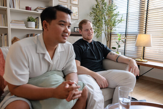 Two men sit on a couch in a bright room.