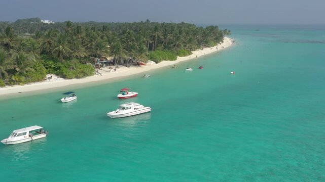 Aerial view of a lively Thoddoo island beach featuring anchored speedboats and jet skis in clear turquoise water, alongside a white sand shore lined with lush palm trees and relaxing tourists.
