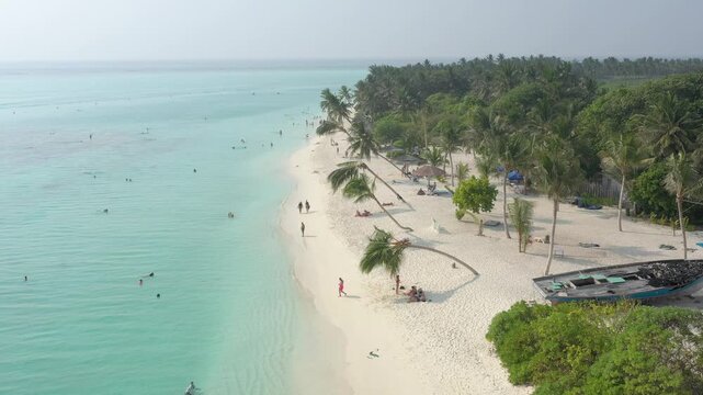 Scenic aerial view along a Thoddoo island beach featuring iconic leaning palm trees, sun umbrellas, a wooden island sign, and tourists enjoying the white sand and clear turquoise ocean.