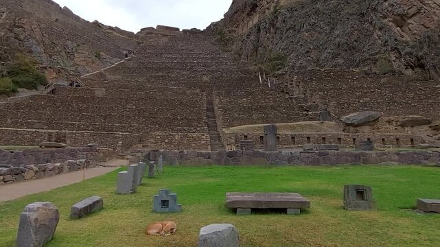 Ollantaytambo, Peru - 12 December 2022. Wide shot of Inca Temple Hill terraces with central staircase, stone retaining walls, lower plaza stones, and rocky Andean slopes under cloud.