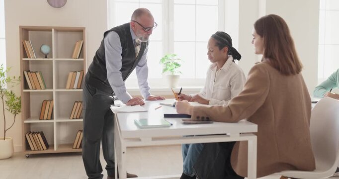 Teacher guides adult students during classroom discussion. Mentor shares guidance while participants take notes at bright desks in a tidy university setting. Strong theme of team education.