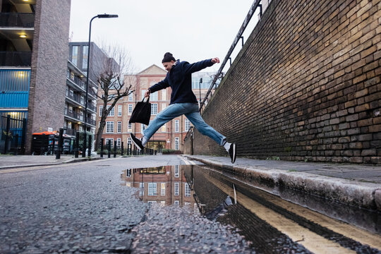 Urban man jumping over street puddle reflecting city buildings