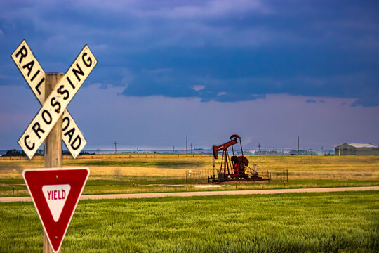 A railroad crossing and yield sign in the foreground with an active oil pump jack in a Kansas field under a moody sky. A blend of transport and energy in the heartland