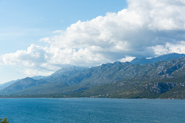 Obraz premium Majestic karst mountain range rising above calm blue Adriatic bay with small coastal settlement and dramatic white clouds. Concept of breathtaking Mediterranean nature and outdoor adventure tourism.