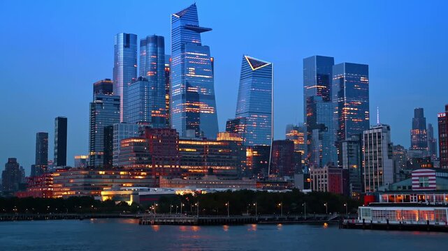 Manhattan skyscrapers and Hudson Yards at twilight hour. High rise glass buildings and urban towers illuminated against a deep blue evening sky.
