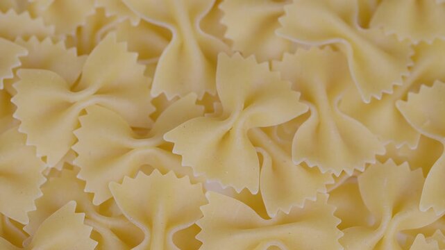 Tightly stacked pale golden farfalle pasta with detailed surface and warm lighting. Close view of unbaked bow shaped pasta with granular texture and gentle pantry ambiance