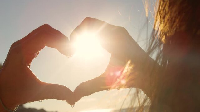 CLOSE UP, LENS FLARE, SILHOUETTE: Unrecognizable woman making a heart shape with her hands, capturing a bright sunburst. Windy hair glows while the sea and beach blur into a warm, golden sunlight.