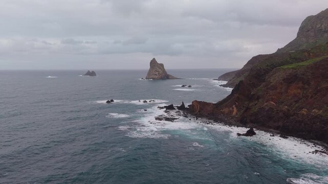 Majestic aerial view of the Atlantic Ocean waves crashing against the rugged volcanic cliffs and Roques de Anaga on a cloudy day. Tenerife.