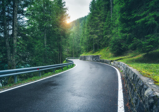 Wet mountain road curving through dense green forest in the Dolomites, Italy. Scenic alpine landscape with asphalt road at sunset, stone wall and guardrail, popular travel and driving route in Alps