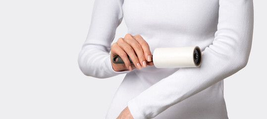 Obraz premium Woman using sticky roller on white long sleeve removing lint, dust on light gray background. Concept of clean outfit maintenance, minimal wardrobe care, polished everyday appearance. Copy space banner