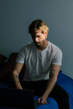 Young man working from home on a laptop, sitting on the edge of a bed