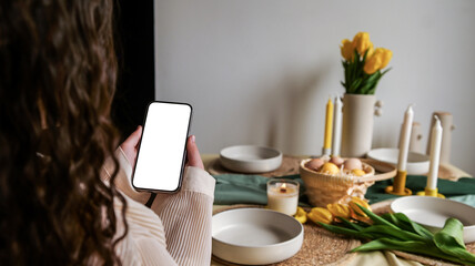 Fototapeta premium Woman holding a smartphone with a blank screen over a festive Easter table setting