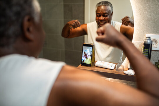 Cheerful senior man flexing during video call in bathroom mirror