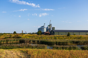 Naklejka premium Golden fields stretch around a serene pond, with a grain storage facility and cranes rising against a clear blue sky, capturing rural life